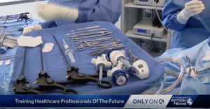 A surgical tray with various medical instruments, including forceps and laparoscopic tools, is displayed in a hospital setting. Two healthcare professionals in blue scrubs and gloves are visible in the background. News banner reads "Training Healthcare Professionals Of The Future.